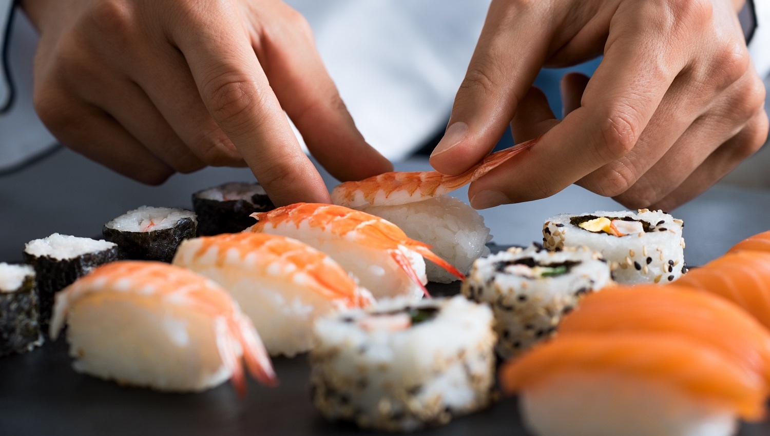 Ein Koch bereitet in der Küche einer Sushi-Bar in Handarbeit Sushi zu. Verschiedene Sushi-Rollen liegen auf dem Tisch.