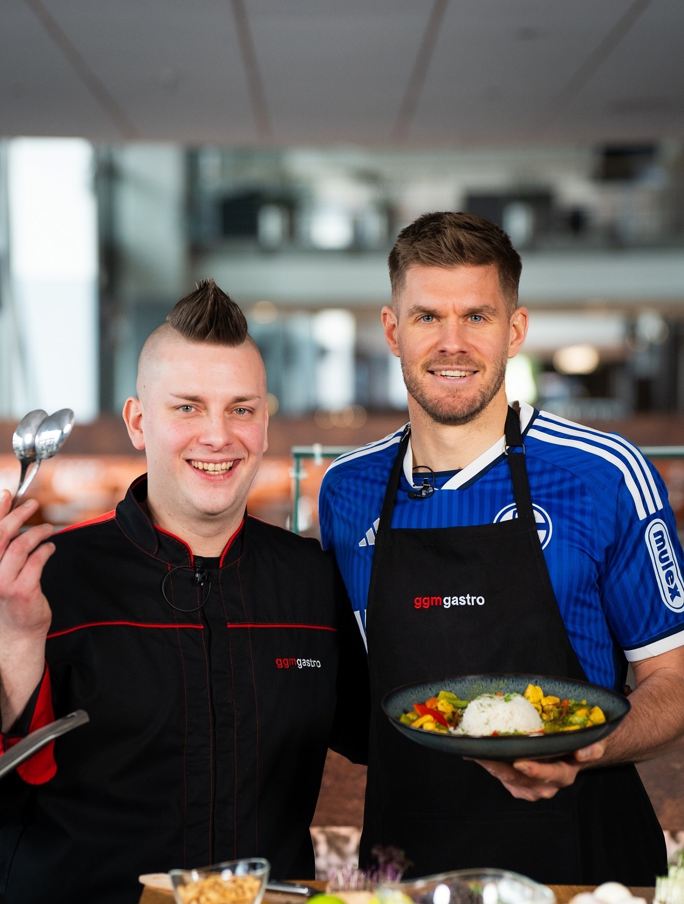 Schalke 04-Stümer Simon Terodde und GGM Gastro-Koch Harry stehen in der Küche. Simon Terodde hält einen Teller Hähnchencurry mit Reis in der Hand, den die beiden nach seinem Rezept zubereitet haben.