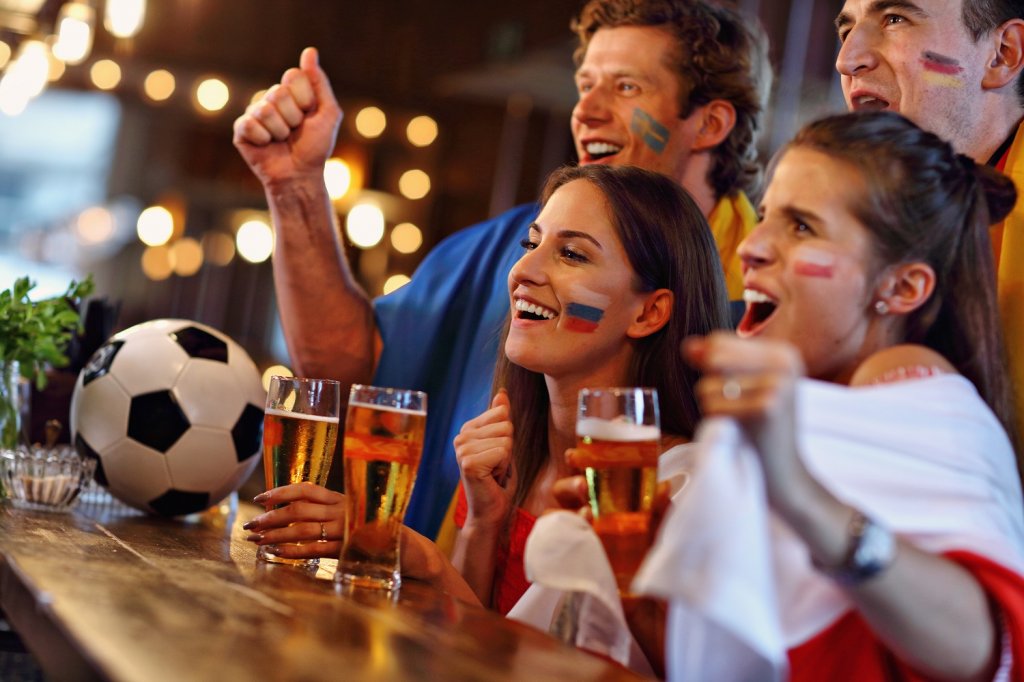 Eine Gruppe Fußballfans trinkt Bier und verfolgt ein Spiel beim Public Viewing in einer Gastronomie. Auf ihren Wangen sind mit Schminke die Flaggen verschiedener Länder gemalt.