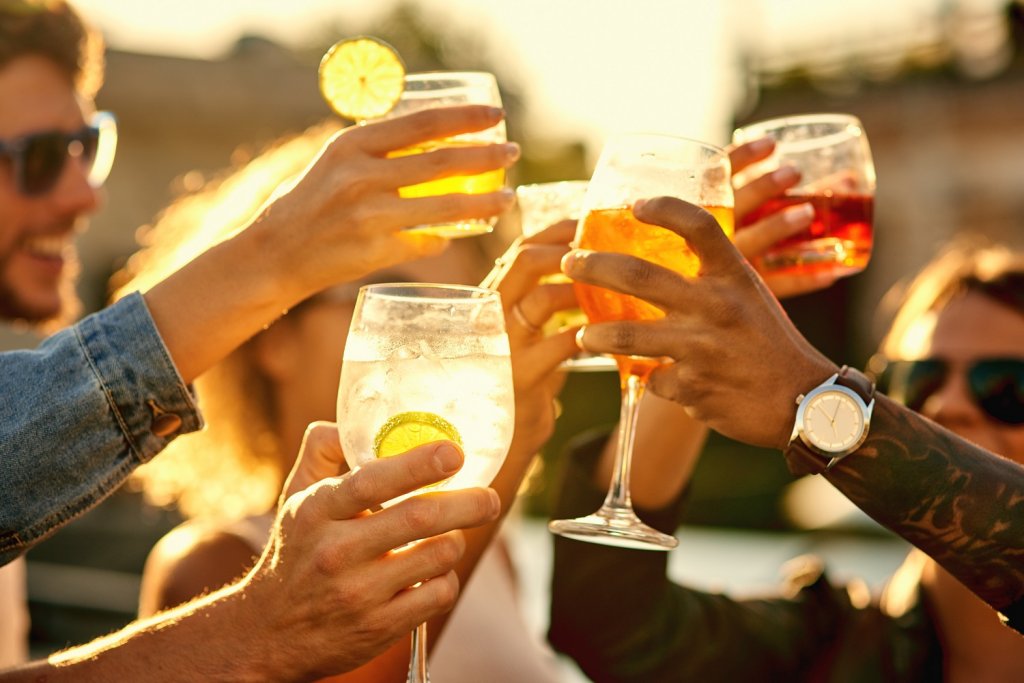 Eine Gruppe an Personen sitzt im Sommer abends an einem Tisch auf der Außenterrasse einer Bar. Die Personen stoßen mit ihren Gläsern an, in den Gläsern sind verschiedene Drinks und Cocktails.
