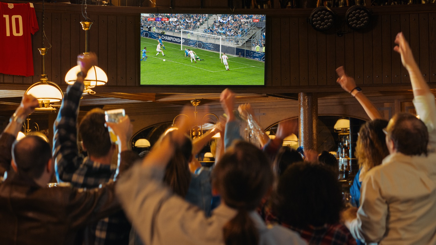 Viele Fußballfans stehen beim Public Viewing zur Europameisterschaft in einer Gastronomie. Sie schauen auf einen großen Fernseher und jubeln für das Team, das gerade ein Tor erzielt.