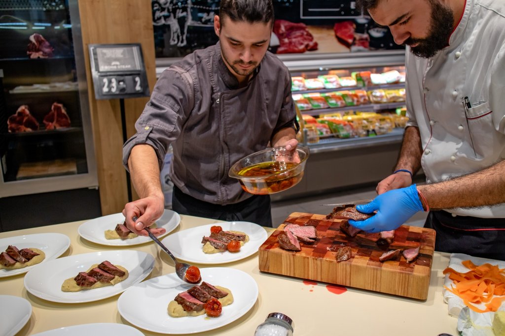 Zwei Köche stehen in einem Restaurant und bereiten ein Gericht mit Scheiben von Dry Aged Ribeye Steak zu. Im Hintergrund ist ein Schrank für Dry Aging zu sehen.