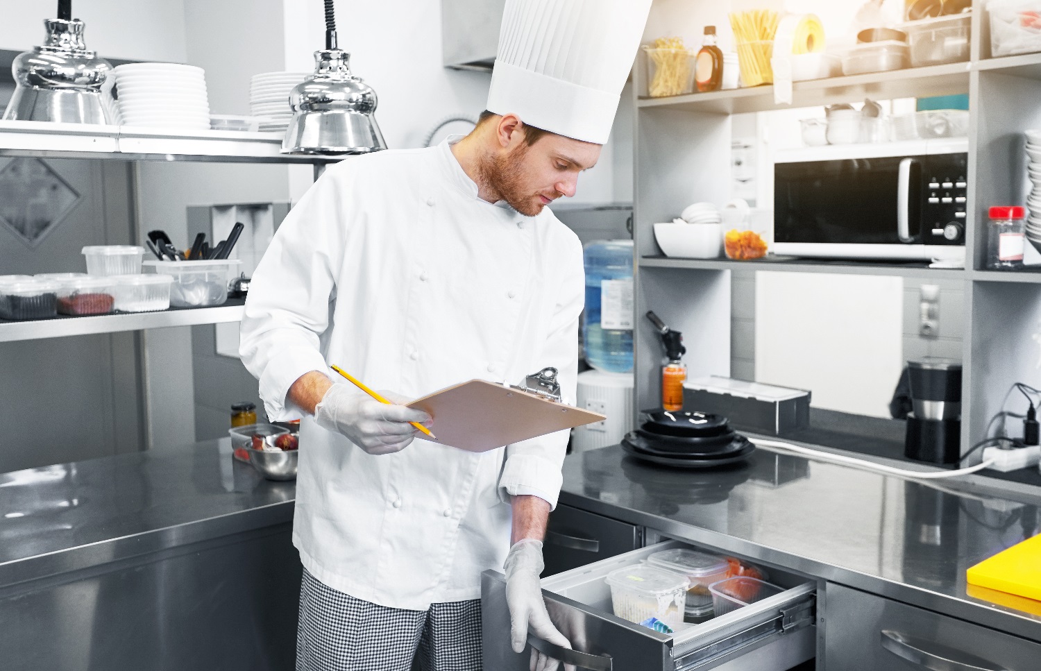 Ein Mitarbeiter in einem Restaurant kontrolliert entsprechend des HACCP-Konzepts die Lebensmittelhygiene in der Küche. Er hält ein Klemmbrett und prüft die Inhalte einer Schublade.