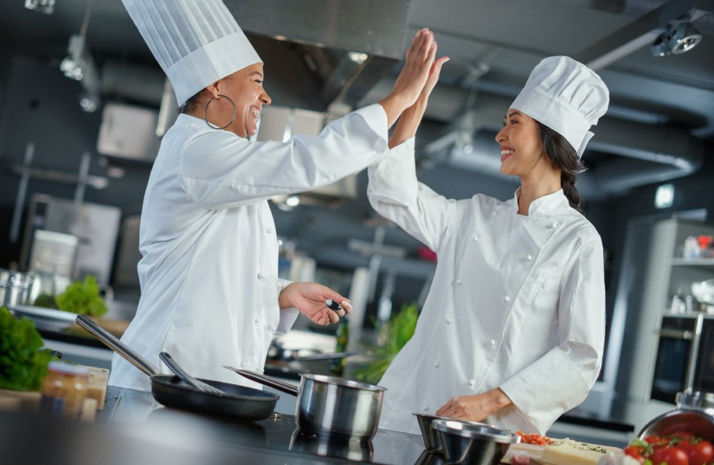 Zwei lächelnde Köchinnen stehen in der Küche eines Restaurants und geben sich ein High Five. Sie stehen symbolisch dafür, dass eine hohe Mitarbeiterzufriedenheit eine bessere Zusammenarbeit ermöglicht.