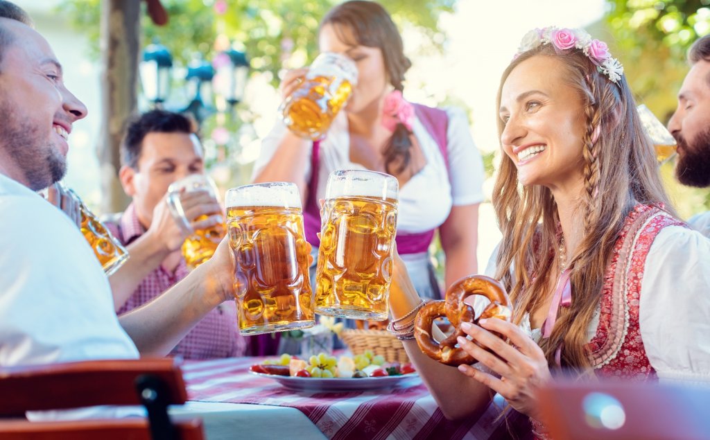 Eine Gruppe an Menschen in bayerischer Tracht sitzt gemeinsam an einem Tisch und stößt mit Bierkrügen an. Außerdem genießen sie traditionelle Oktoberfest Gerichte wie Brezn.