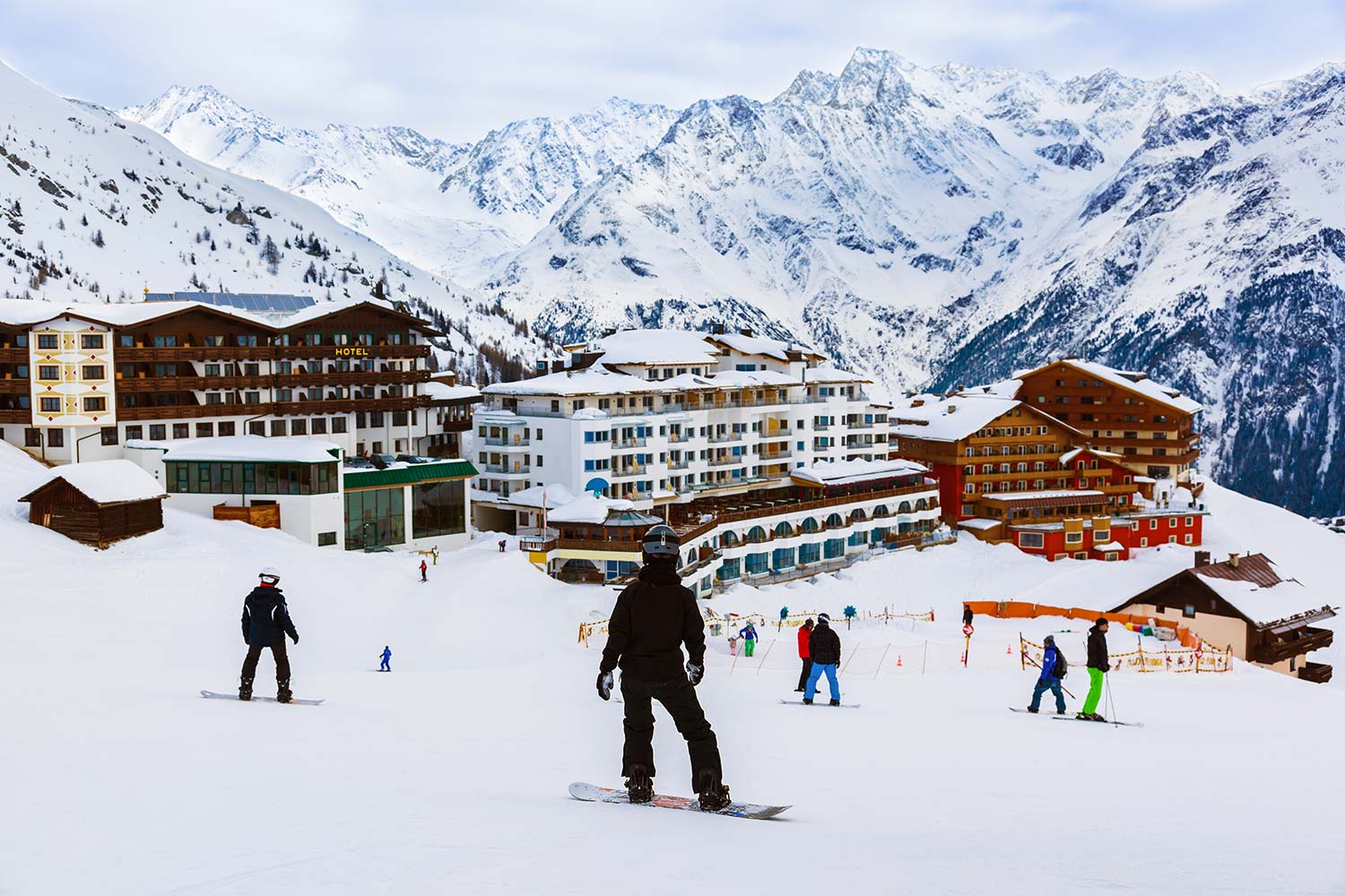 Das Bild zeigt ein verschneites Skigebiet in den Bergen. Im Vordergrund sind Wintersport-Touristen auf Snowboards und Skiern zu sehen. Im Hintergrund befinden sich mehrere malerische Hotels.