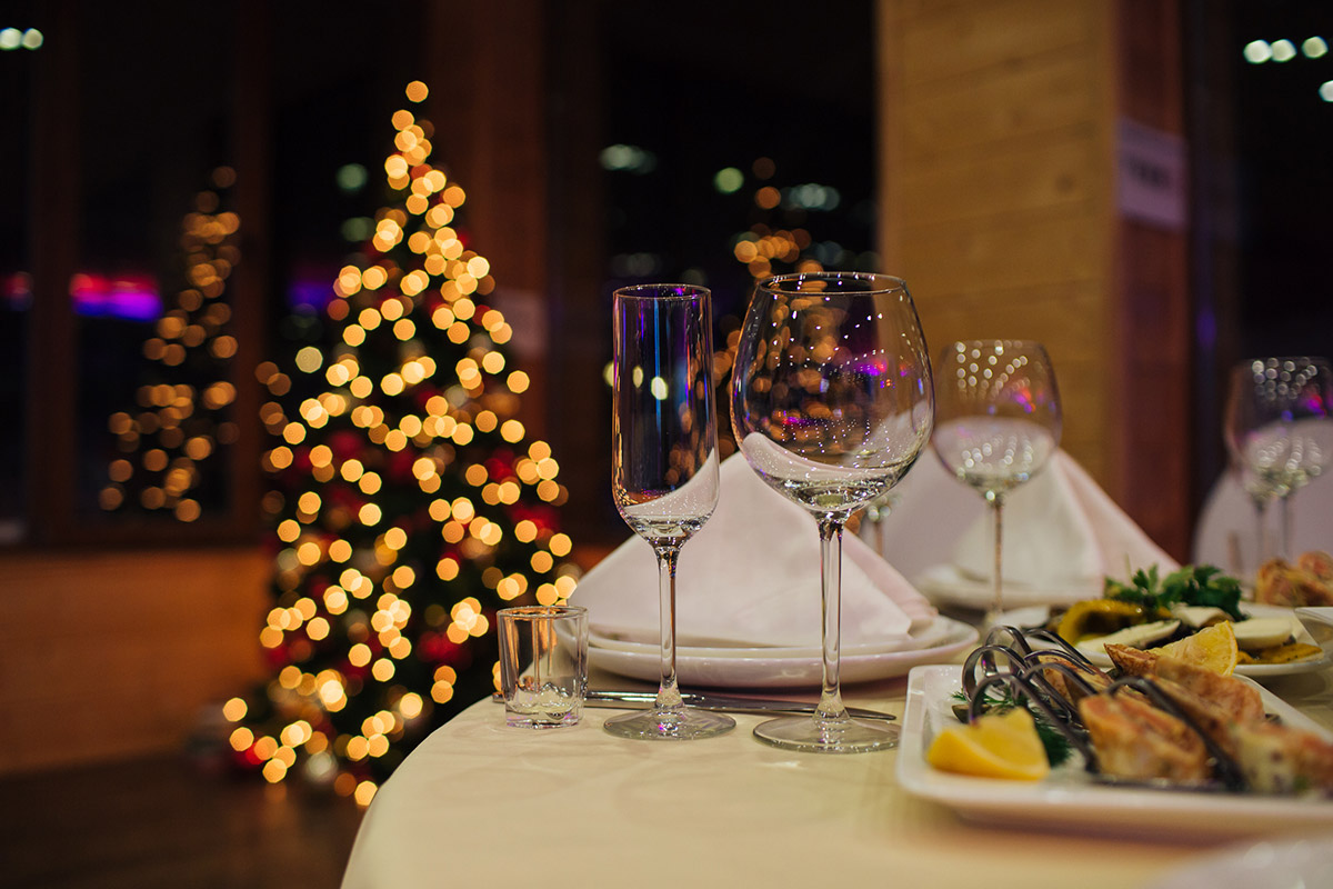 Weihnachtsfeier Catering in einem Restaurant: Im Vordergrund ein gedeckter Tisch mit verschiedenen Gläsern und Essen auf Tellern, im Hintergrund festliche Dekoration und Weihnachtsbäume mit Lichterketten.