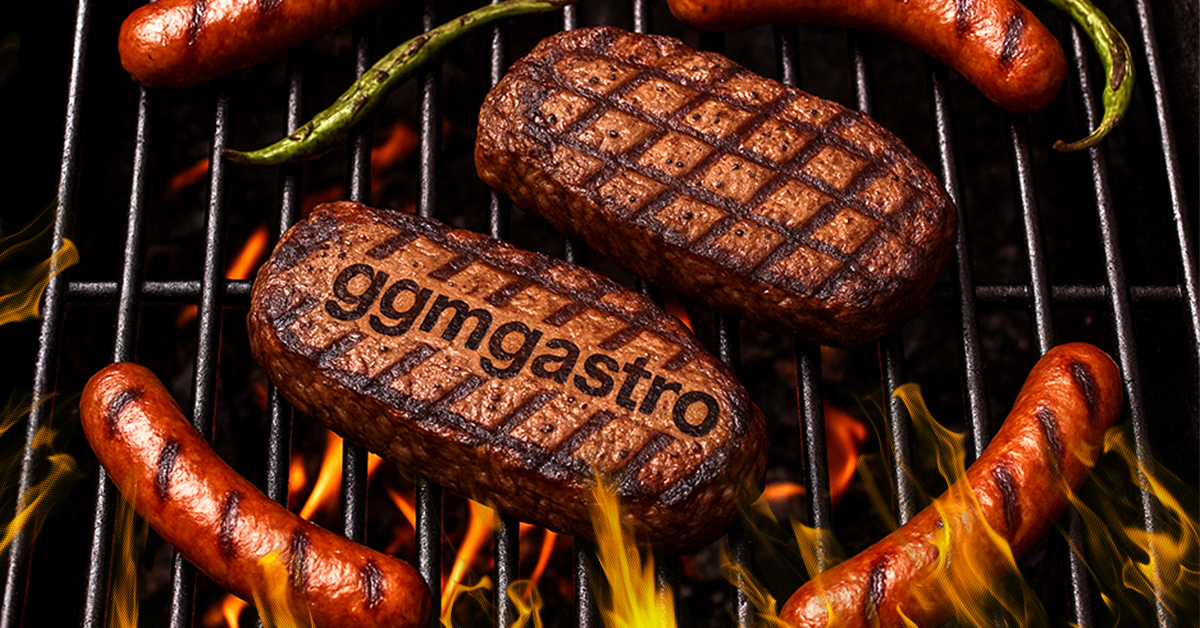 Zwei Steaks mit Grillmuster, eines davon mit dem eingebrannten "ggmgastro"-Schriftzug, liegen neben Bratwürstchen und Pimientos auf einem Grillrost über einem Kohlenfeuer.